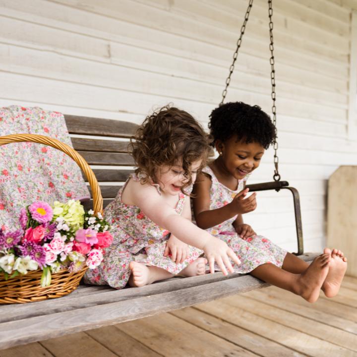 Baby and Toddler sitting on a porch swing wearing Rowena Gauze Dress next to a printed blanket and basket of flowers.