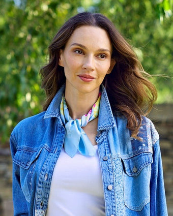 Tulip Scarf tied around neck on a model wearing a denim shirt, jeans and white tank top standing outside under greenery.